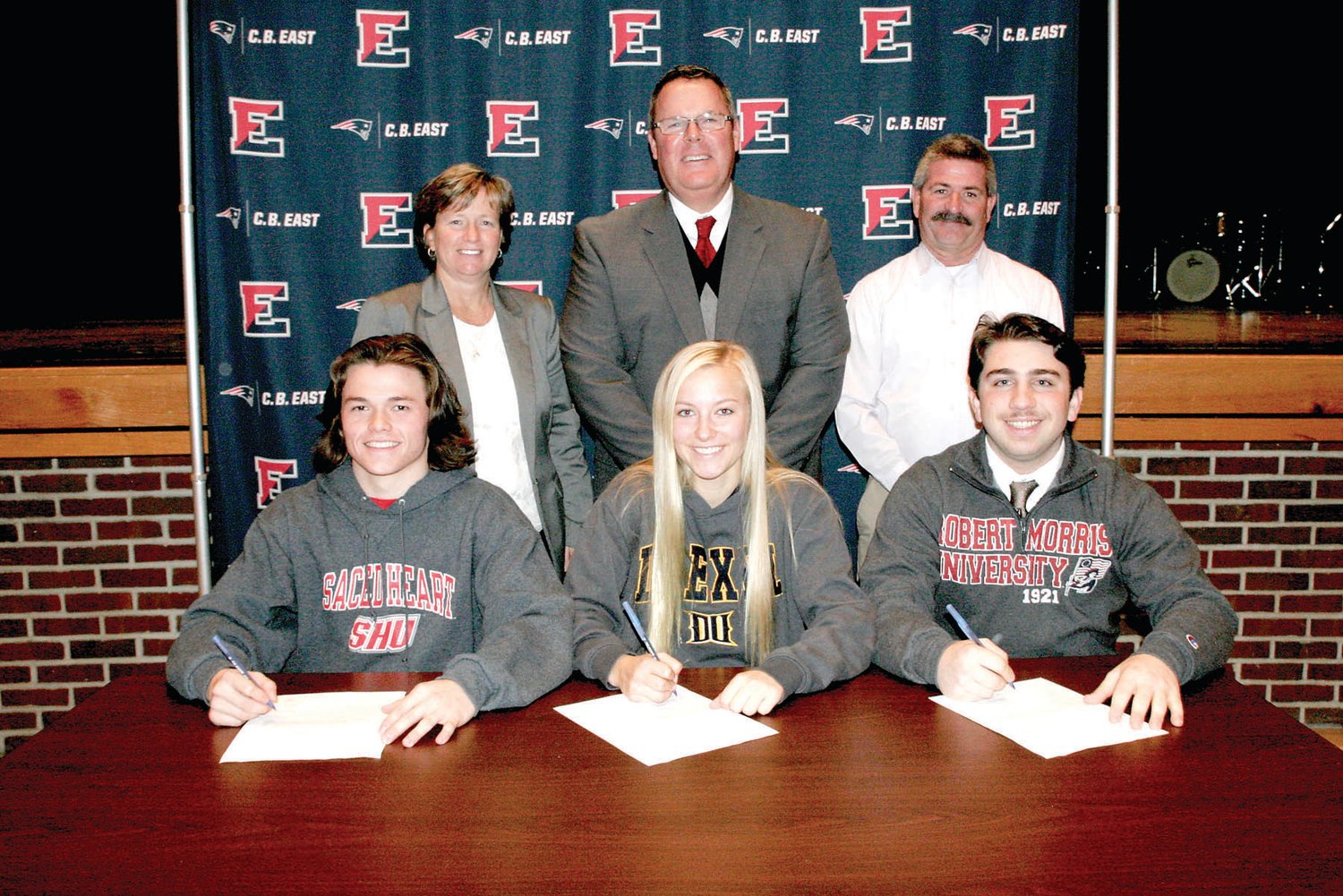 CB East seniors, from left, front row, Ryan Stout, Cailey Lever and Liam Rosenthal sign letters of intent to play at Sacred Heart, Drexel and Robert Morris universities, respectively. Joining the trio at the signing ceremony are, from left, back row, Principal Lori Gallagher-Landis, Class of 2019 House Principal Luke Hadfield and Athletic Director John Reading. Photograph by Mary Jane Souder