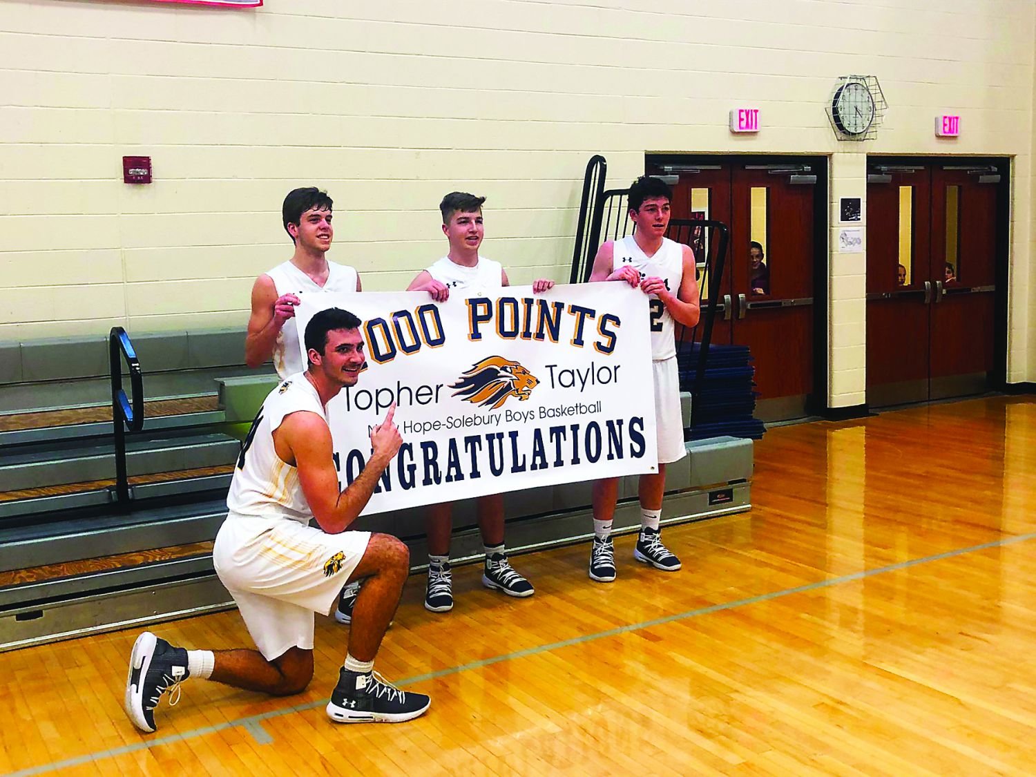 New Hope-Solebury senior Topher Taylor, kneeling, tallied his 1,000th career point March 9 in the Lions’ first state playoff win under longtime head coach Rick Fedele. Holding the celebration banner are, from left, seniors Connor Wallace, Logan Waterman and Pat Cooney.