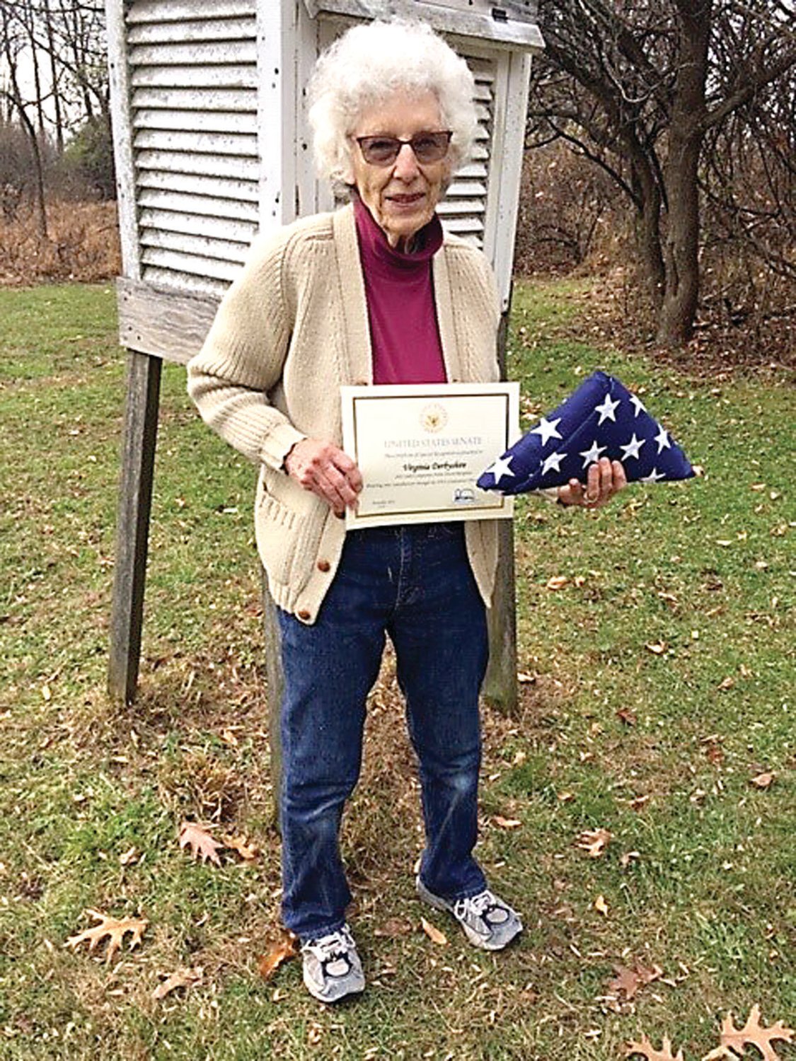 Virginia Derbyshire, standing in front of her weather station, displays her national NOAA award for outstanding volunteer service and an America flag that flew over the U.S. Capitol.
